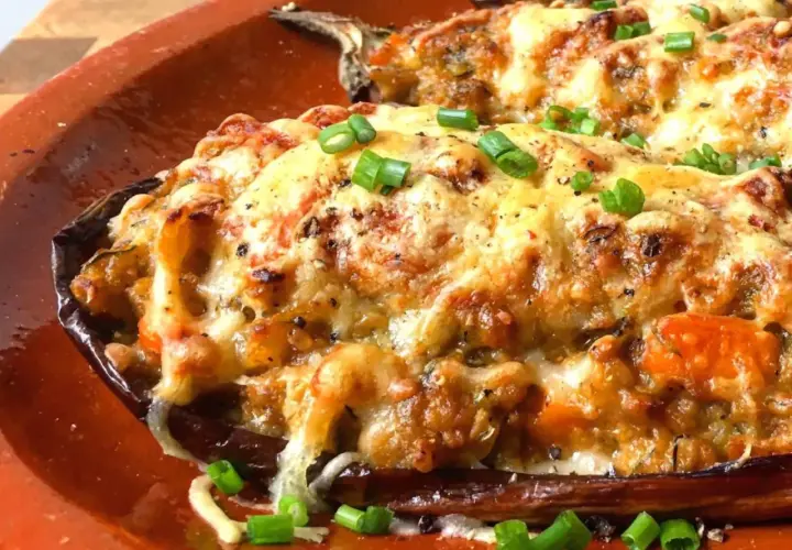 Berenjenas rellenas gratinadas con queso derretido y cebolleta picada, servidas en una cazuela de barro.