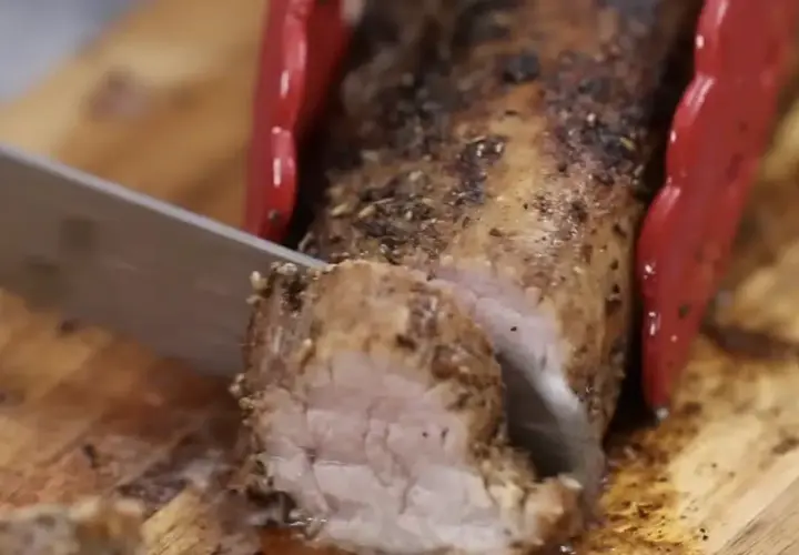 Un jugoso solomillo de cerdo al horno, recién cortado en una tabla de madera. Las pinzas rojas sujetan la carne mientras un cuchillo la rebana.
