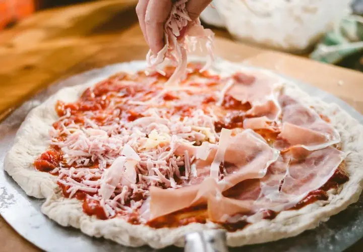 Manos esparciendo queso rallado sobre una pizza casera con jamón y salsa de tomate.