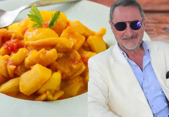 Un hombre con gafas de sol y traje blanco sonríe junto a un plato de papas con choco, un guiso amarillo con trozos de tomate y perejil.