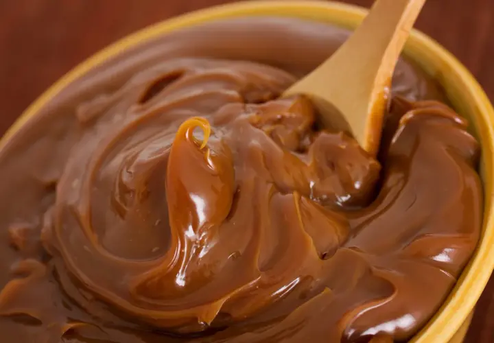 Cuchara de madera removiendo un tazón lleno de dulce de leche cremoso, perfecto para tu dulce de leche receta.