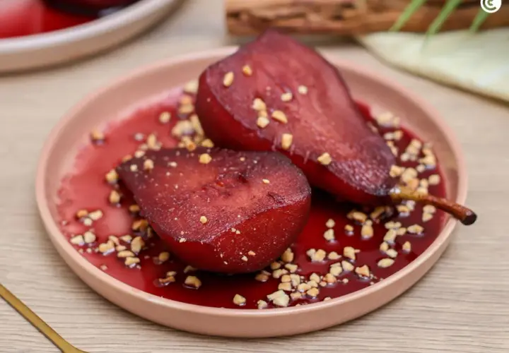 Peras al vino, tiernas y brillantes, servidas en un plato rosa con trocitos de nuez y un sirope rojo intenso.