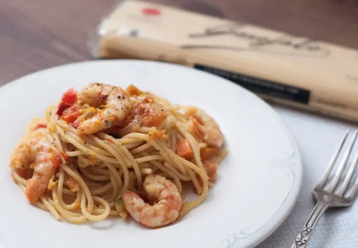 Espaguetis con gambas y trocitos de tomate, servidos en un plato blanco. Un tenedor plateado descansa a la derecha.