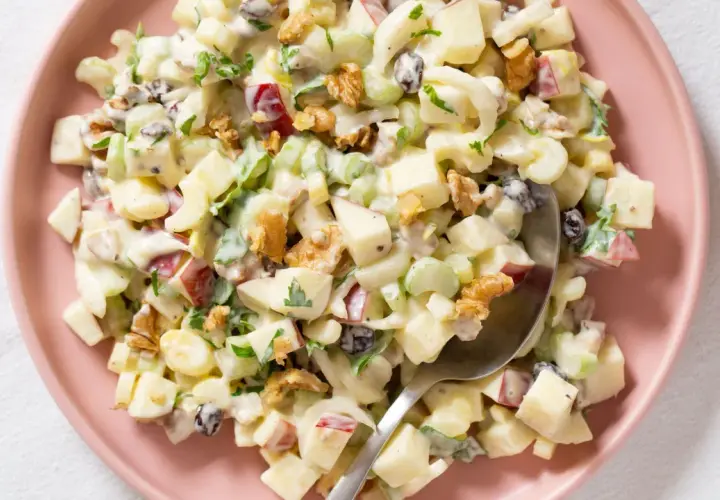 Ensalada Waldorf cremosa con trozos de manzana, apio, nueces y pasas, servida en un plato rosa.