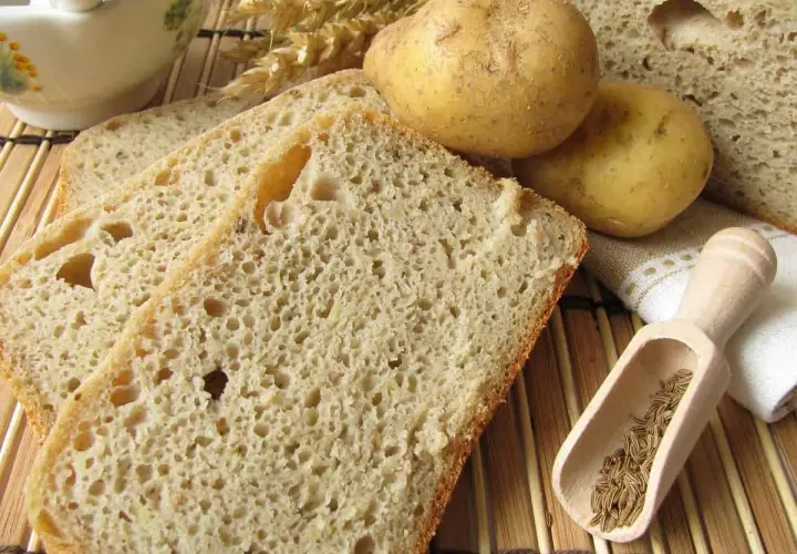 Rebanadas de pan de patata rústico, con patatas enteras, espigas de trigo y comino en una cuchara de madera.