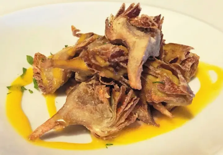 Montón de alcachofas fritas crujientes, doradas y sazonadas, servidas sobre un plato blanco con un toque de salsa amarilla y perejil picado.