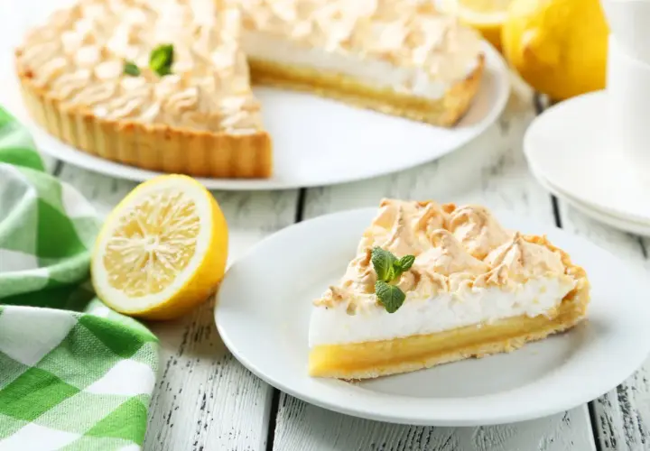 Delicioso pastel de limón con merengue dorado, decorado con menta fresca. Un trozo servido en plato blanco, junto a limones y una taza de té.
