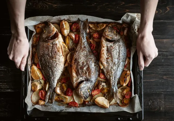 Tres pescados dorados al horno, acompañados de rodajas de limón, tomates cherry y ajos, listos para servir.