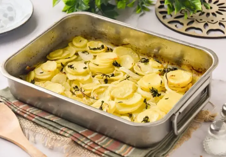 Patatas a lo pobre recién horneadas en una bandeja metálica, con rodajas finas de patata y cebolla doradas y hierbas aromáticas.