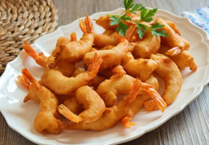 Un plato blanco lleno de gambas a la gabardina doradas y crujientes, decorado con perejil fresco.