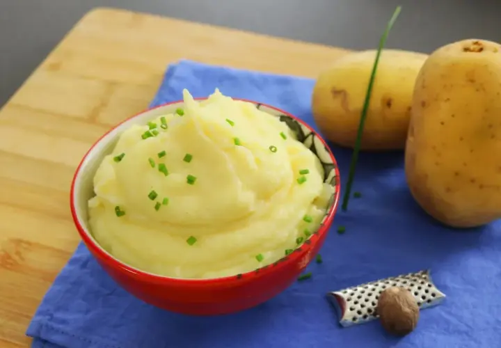 Un cremoso puré de patatas, adornado con cebollino fresco, junto a patatas crudas y una nuez moscada.