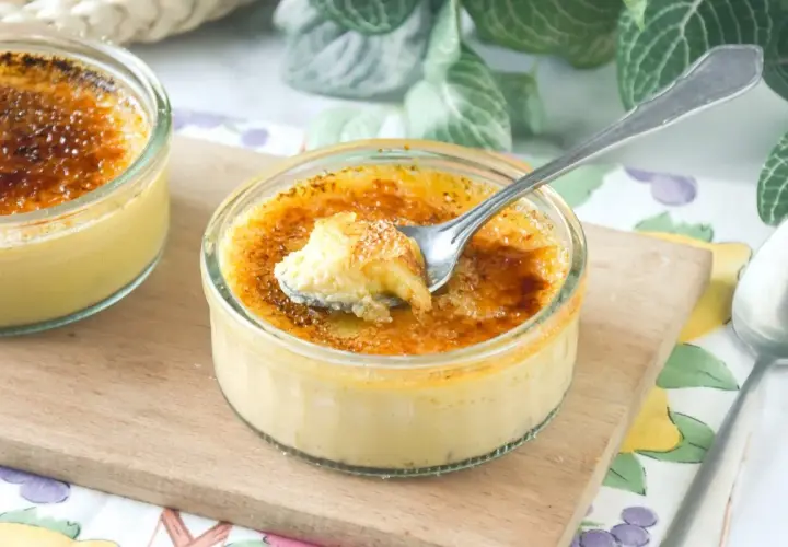 Dos cremosos postres de creme brulee en vasitos de cristal, uno con una cucharada sacada, sobre una tabla de madera.
