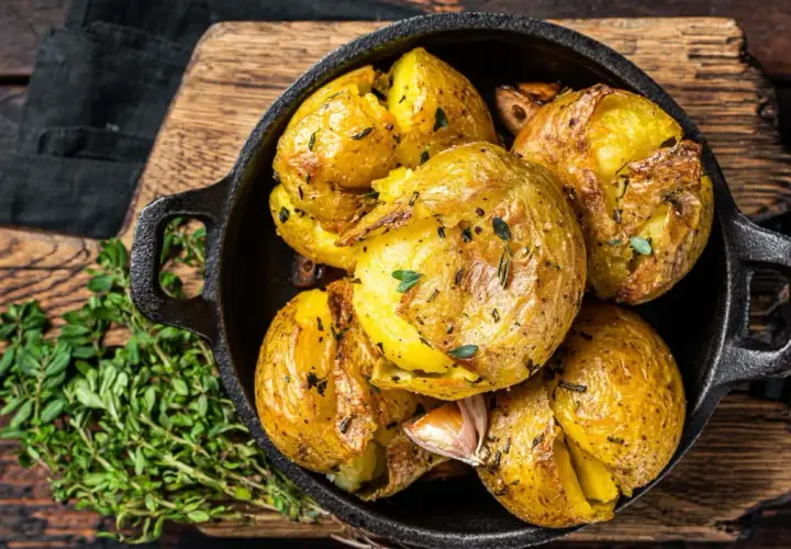 Patatas al horno doradas y crujientes, con hierbas y ajo, servidas en una sartén de hierro fundido sobre una tabla de madera.