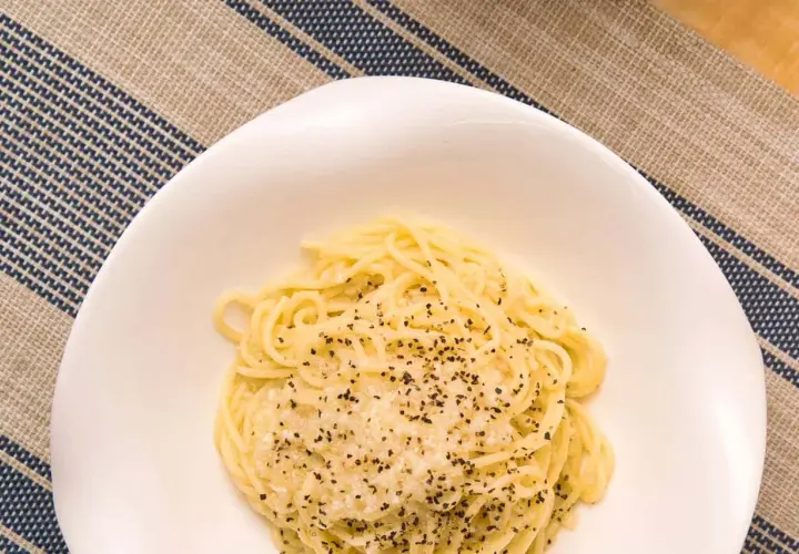 Un plato de cacio e pepe, pasta cremosa con queso y pimienta negra, servido en un cuenco blanco sobre un mantel a rayas.
