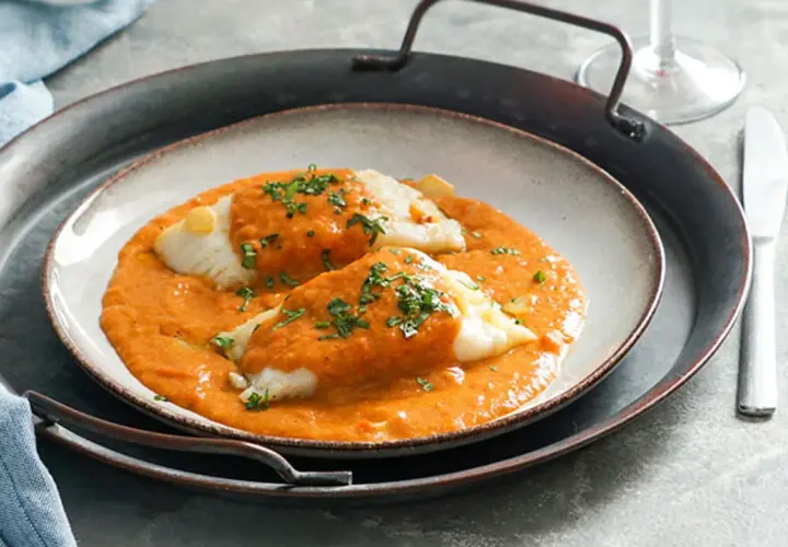 Dos lomos de bacalao a la vizcaína, cubiertos de salsa roja y perejil picado, servidos en un plato sobre una bandeja metálica.