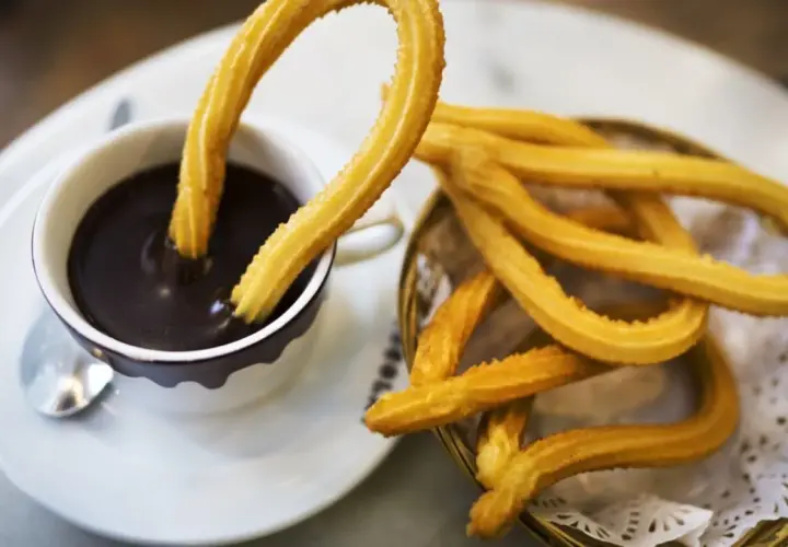 Churros sin gluten dorados y crujientes, uno sumergido en una taza de chocolate caliente espeso.