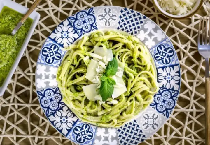 Un plato de pasta al pesto, decorado con queso parmesano y albahaca fresca, junto a una pequeña porción de salsa pesto.