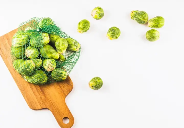 Una bolsa de malla verde llena de coles de Bruselas frescas descansa sobre una tabla de cortar de madera. Algunas coles de Bruselas están esparcidas alrededor.