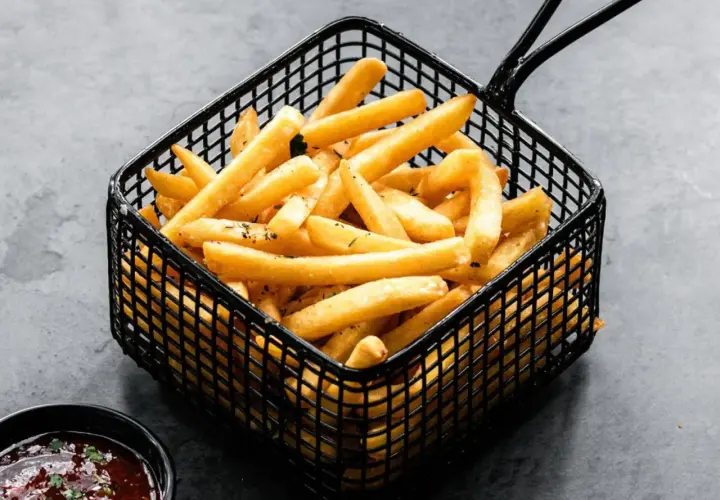 Cesta negra llena de crujientes patatas fritas doradas, con un cuenco de salsa roja al lado.