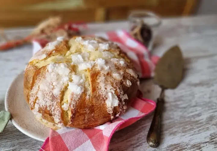 Un pan quemado dorado y crujiente, espolvoreado con azúcar, descansa sobre un plato. Una servilleta a cuadros rojos y blancos y un utensilio para pasteles completan la escena.