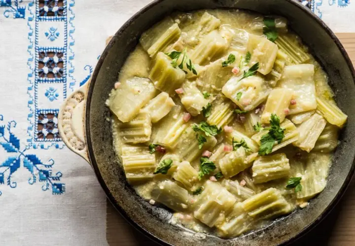 Cardo en salsa cremosa con trocitos de jamón y perejil, servido en sartén sobre mantel bordado. Un plato delicioso con cardo y almendras.