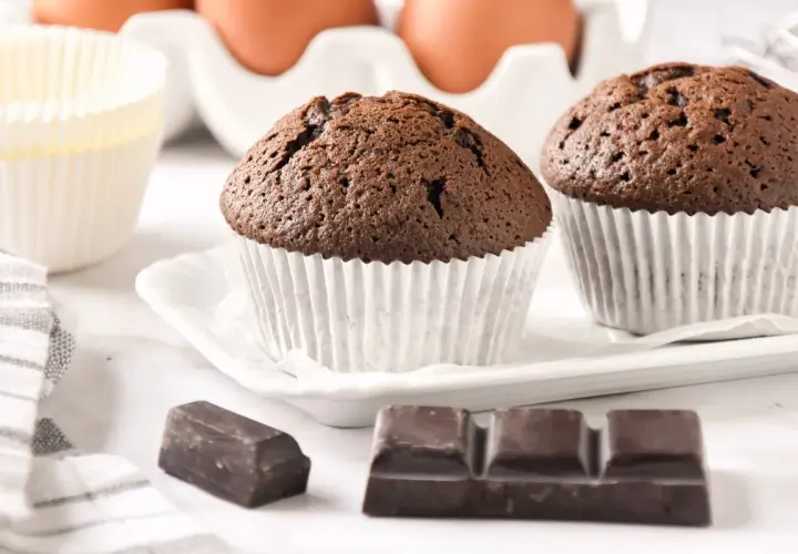 Dos magdalenas de chocolate recién horneadas, listas para disfrutar. A su lado, trozos de chocolate y moldes para hornear.