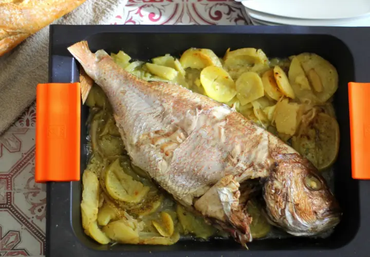 Pargo al horno con patatas y cebolla en una bandeja negra. Un pan rústico descansa a un lado.