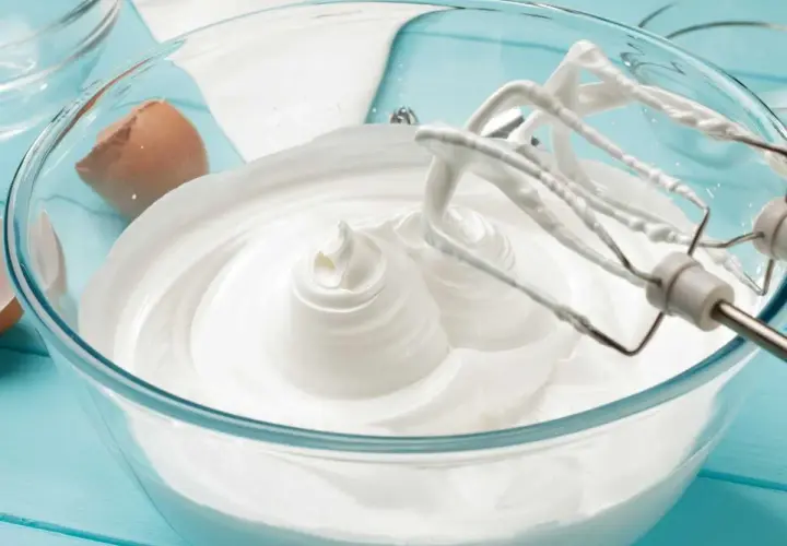 Batidora mezclando merengue italiano en un bol de cristal. Cáscara de huevo al lado.