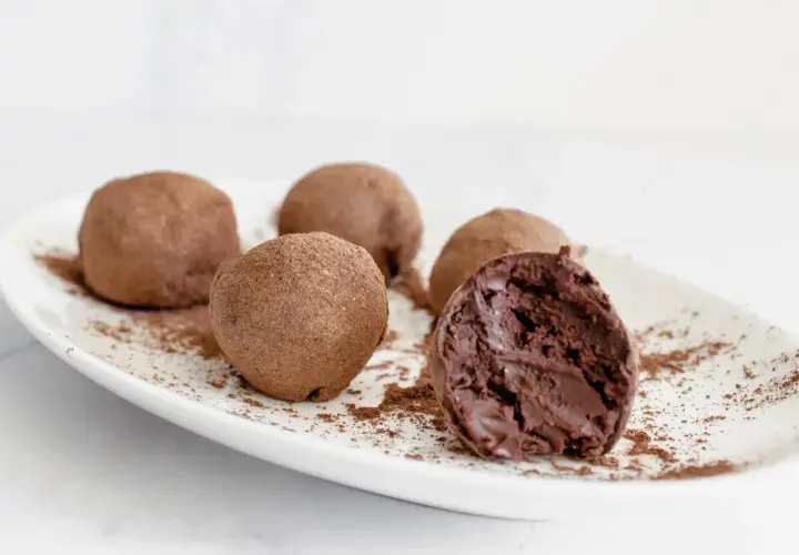 Cinco trufas de chocolate espolvoreadas con cacao en polvo sobre un plato blanco. Una trufa está partida, revelando su interior cremoso.