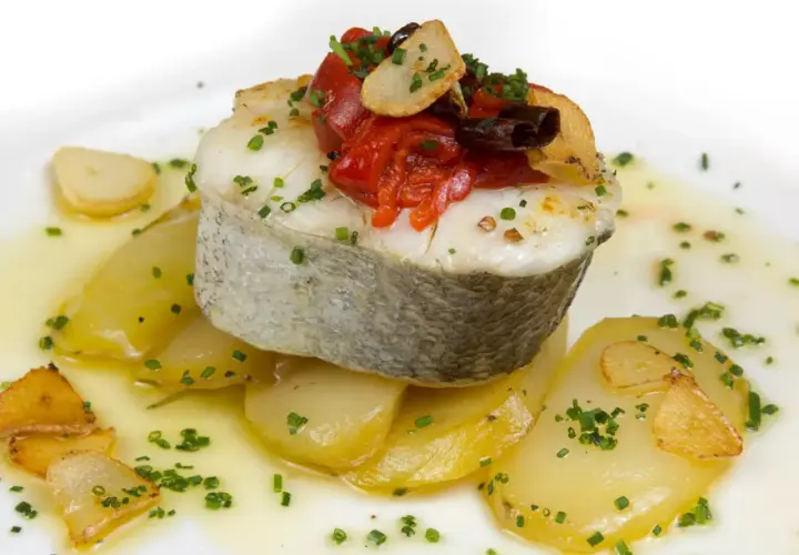 Deliciosa merluza a la plancha sobre patatas, con pimientos asados, ajo crujiente y perejil picado.