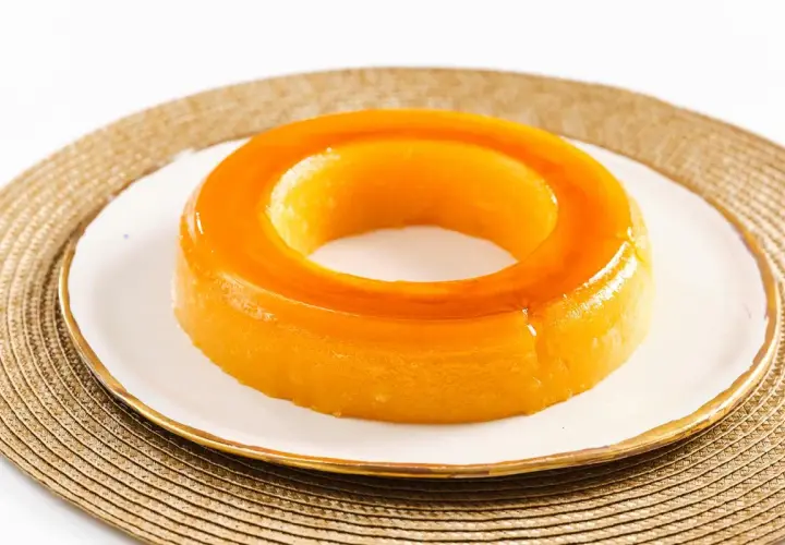 Un delicioso tocino de cielo, con su característico color dorado y forma de anillo, reposa sobre un plato blanco con borde dorado.
