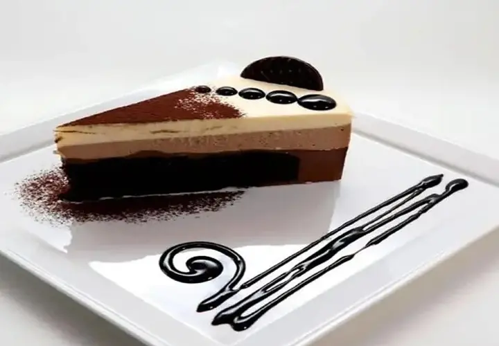 Una rebanada de tarta de tres chocolates, con capas de chocolate blanco, con leche y negro, decorada con cacao en polvo y un disco de chocolate.