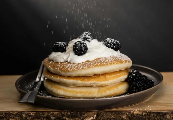Una pila de tortitas americanas cubiertas con crema batida y moras, espolvoreadas con azúcar glas.