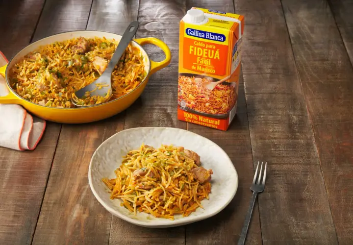 Fideos a la cazuela servidos en un plato y una paellera amarilla, junto a un caldo para fideuá.