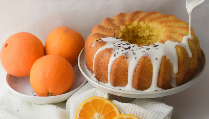 Bizcocho de naranja y yogur con glaseado blanco y chispas de chocolate. Naranjas frescas y rodajas de naranja completan la escena.