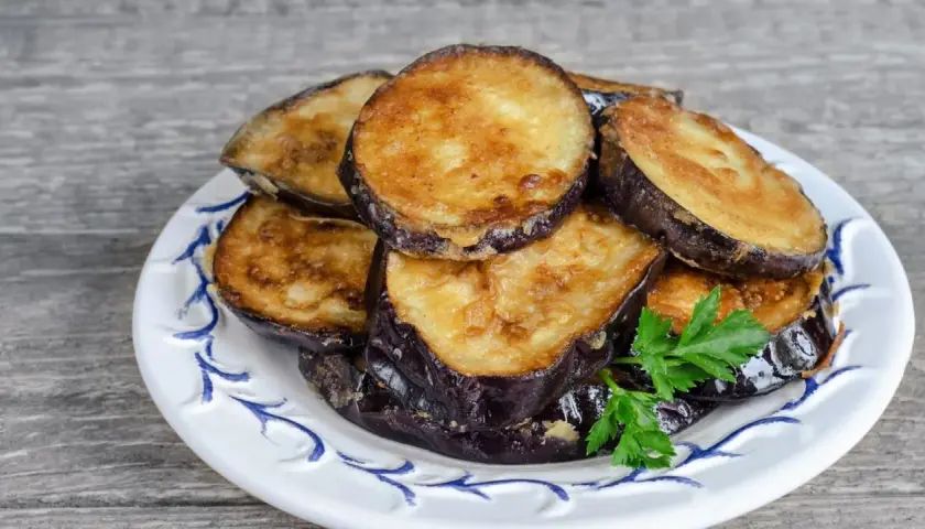 Rodajas doradas de berenjenas fritas apiladas en un plato blanco con decoración azul, adornadas con perejil fresco.