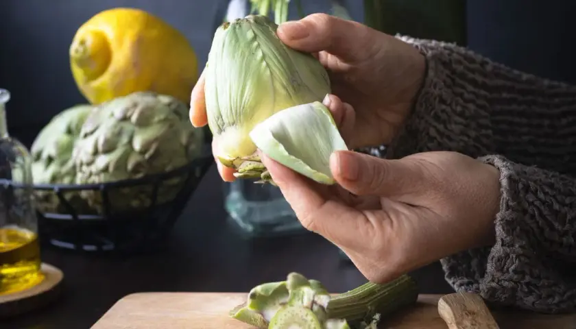 Manos preparan alcachofas para deliciosas recetas. Limón y aceite de oliva a la vista.