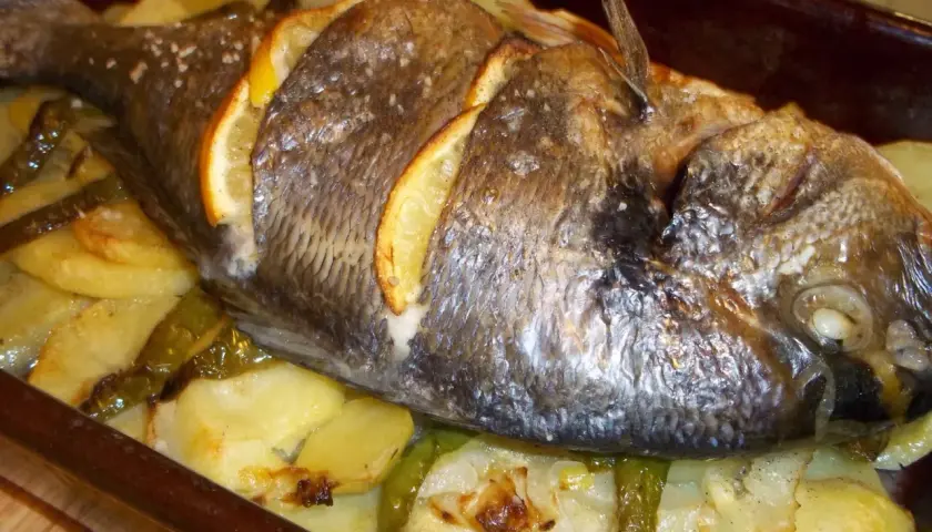 Dorada al horno con rodajas de limón, patatas y pimientos verdes en una bandeja.