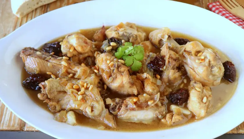 Delicioso pollo guisado con ciruelas pasas y piñones, servido en un plato blanco con pan rústico al fondo.