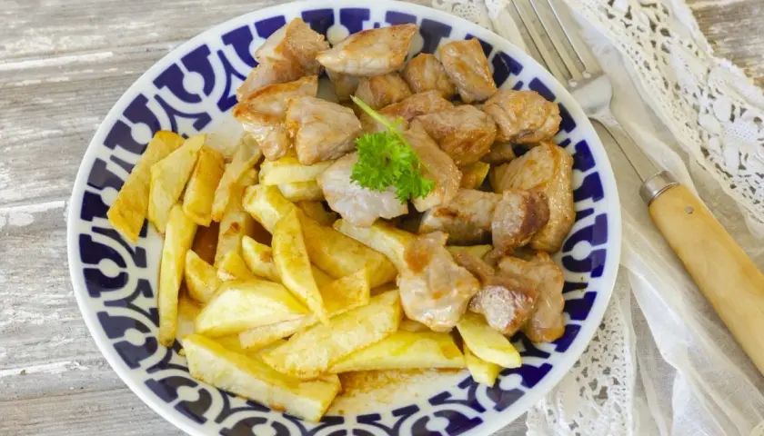 Raxo gallego jugoso con patatas fritas crujientes, servido en un plato con borde azul y blanco.