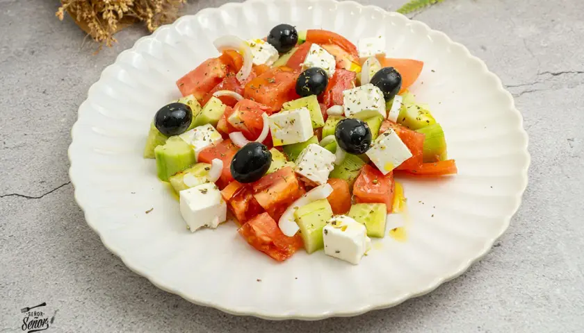 Ensalada griega fresca con tomates, pepinos, cebolla, aceitunas negras y queso feta en cubos, aderezada con aceite de oliva y hierbas.