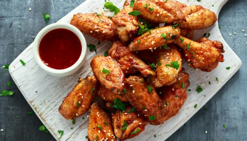Alitas de pollo al horno crujientes, doradas y espolvoreadas con sésamo y perejil, acompañadas de una salsa roja.