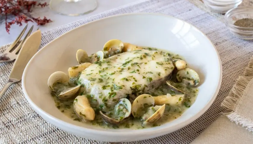 Delicioso pescado en salsa verde con almejas, servido en un plato blanco sobre un mantel de lino.