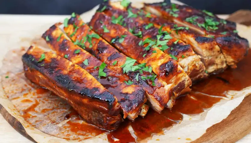 Costillas a la barbacoa jugosas y glaseadas, cubiertas con perejil fresco picado, servidas sobre papel de hornear en una tabla de madera.