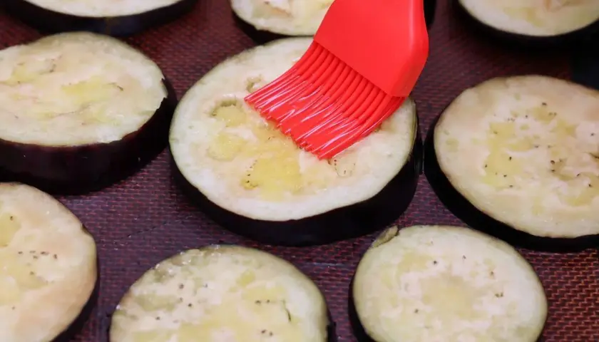 Rodajas de berenjenas listas para ser horneadas, untadas con aceite y especias con un pincel rojo.