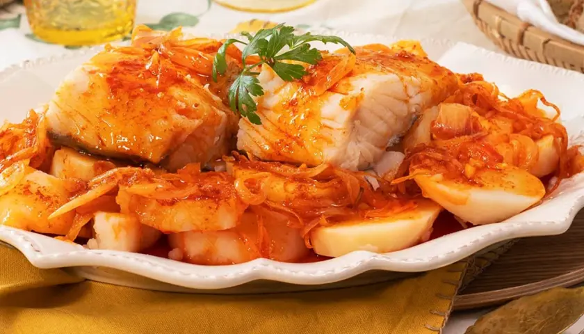 Bacalao a la gallega, un plato tradicional con trozos de pescado blanco cubiertos de una salsa rojiza con cebolla y patatas cocidas.
