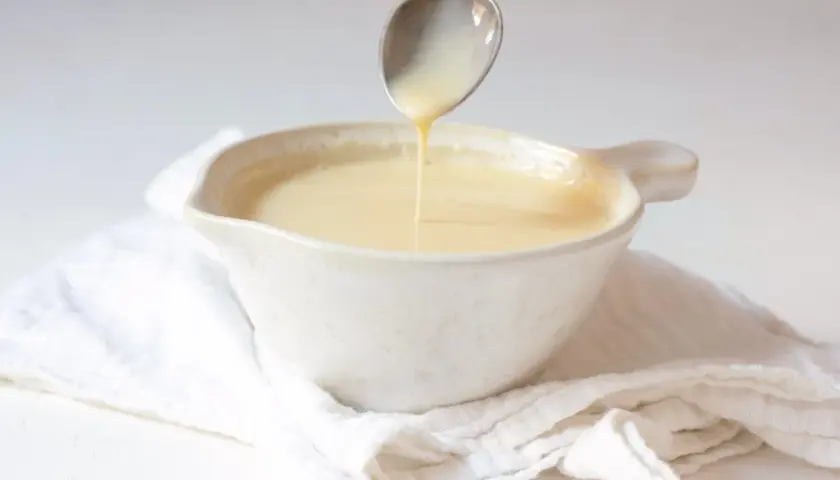 Cuchara vertiendo crema inglesa en un cuenco blanco sobre tela arrugada.