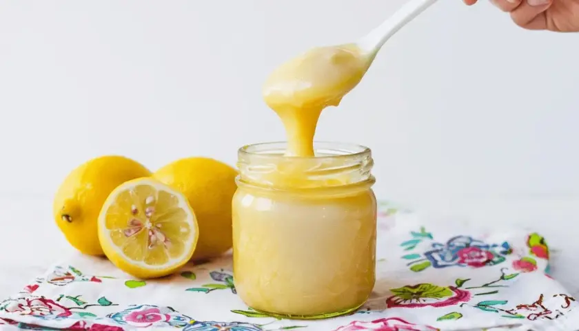 Cucharada de cremoso lemon curd cayendo en un frasco, junto a limones frescos sobre un paño floral.