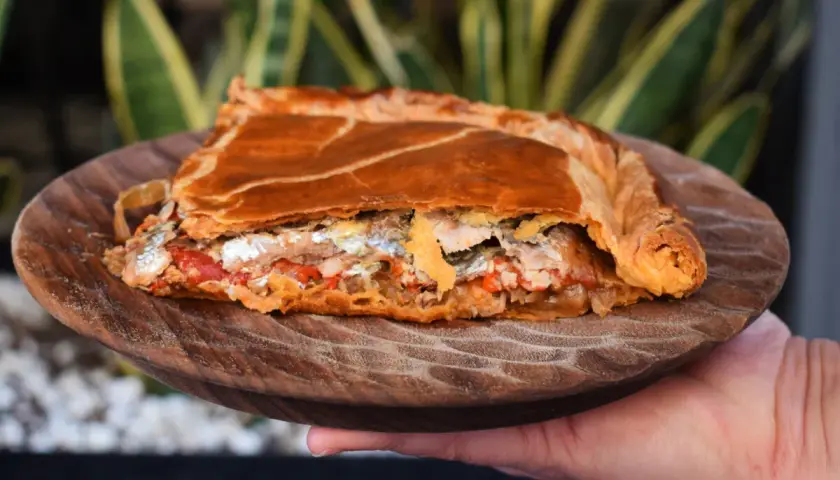 Una porción de empanada gallega, dorada y rellena de pescado y verduras, servida en plato de madera.