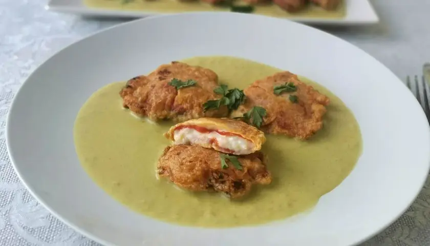 Pimientos rellenos de bacalao rebozados, servidos en una cremosa salsa verde. Uno está cortado, mostrando el relleno blanco y rojo.